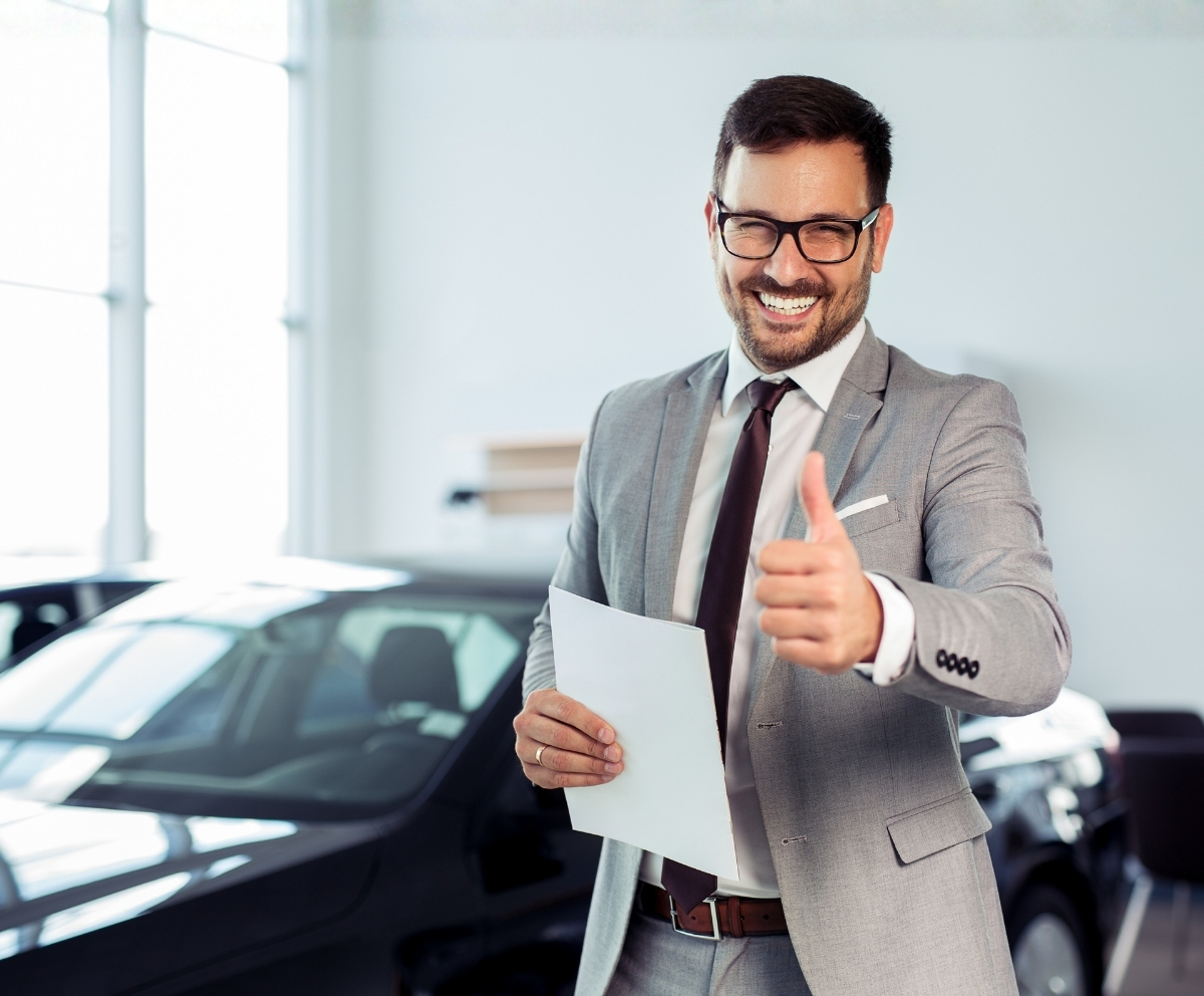 Smiling car dealership representative holding paperwork and giving a thumbs up